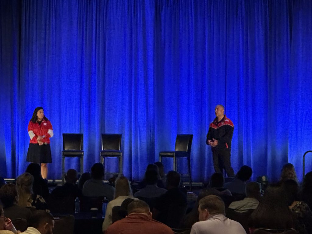 Two presenters on stage in front of blue drapes at a corporate event