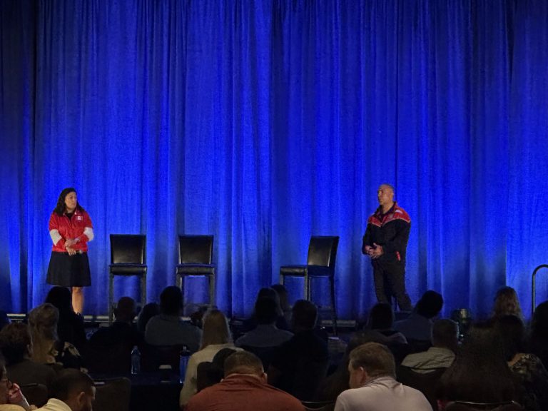 Two presenters on stage in front of blue drapes at a corporate event