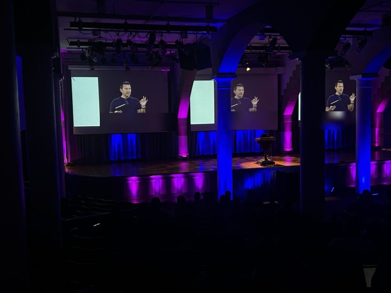Keynote speaker on triple projection screens in a historic hall with purple and blue lighting