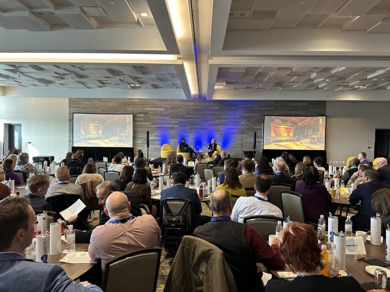 Conference audience in a ballroom with two projection screens and a panel on stage